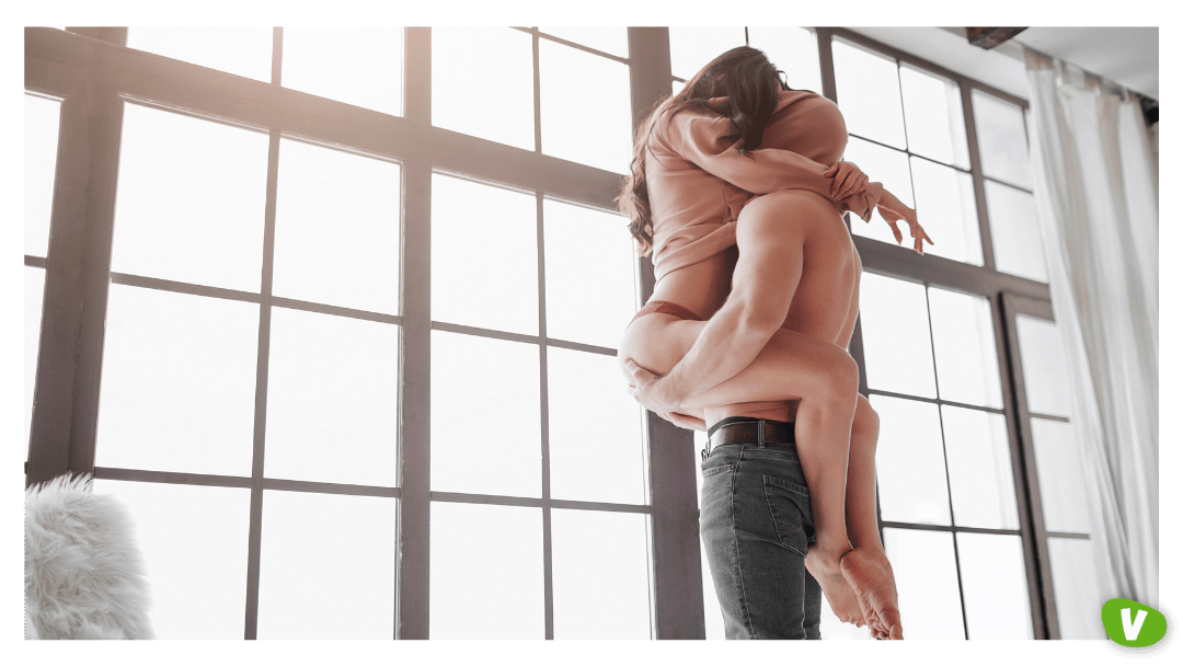 Sexy Couple Stand at Window and Hide Head under Sweater. Guy Hold Woman on Hands. She Sit on Him and Embrace His Body with Legs.